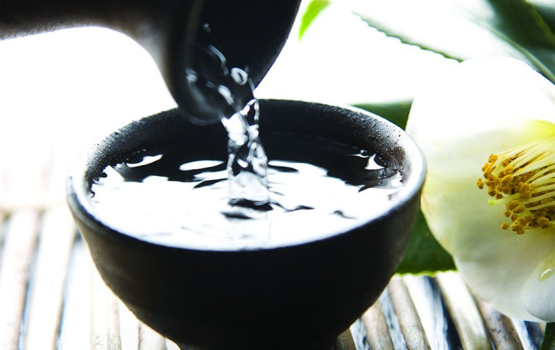 Someone pours a small cup of sake, nestled on a bammboo mat next to a hibiscus flower.